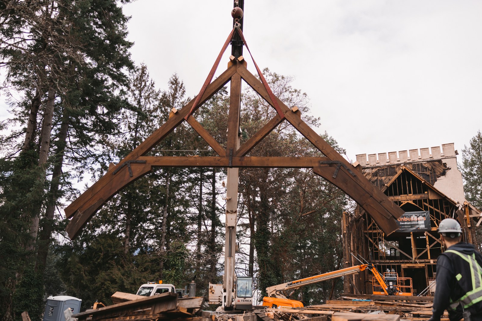 A crane lifts a triangular timber roof frame while a worker watches at an Unbuilders deconstruction site