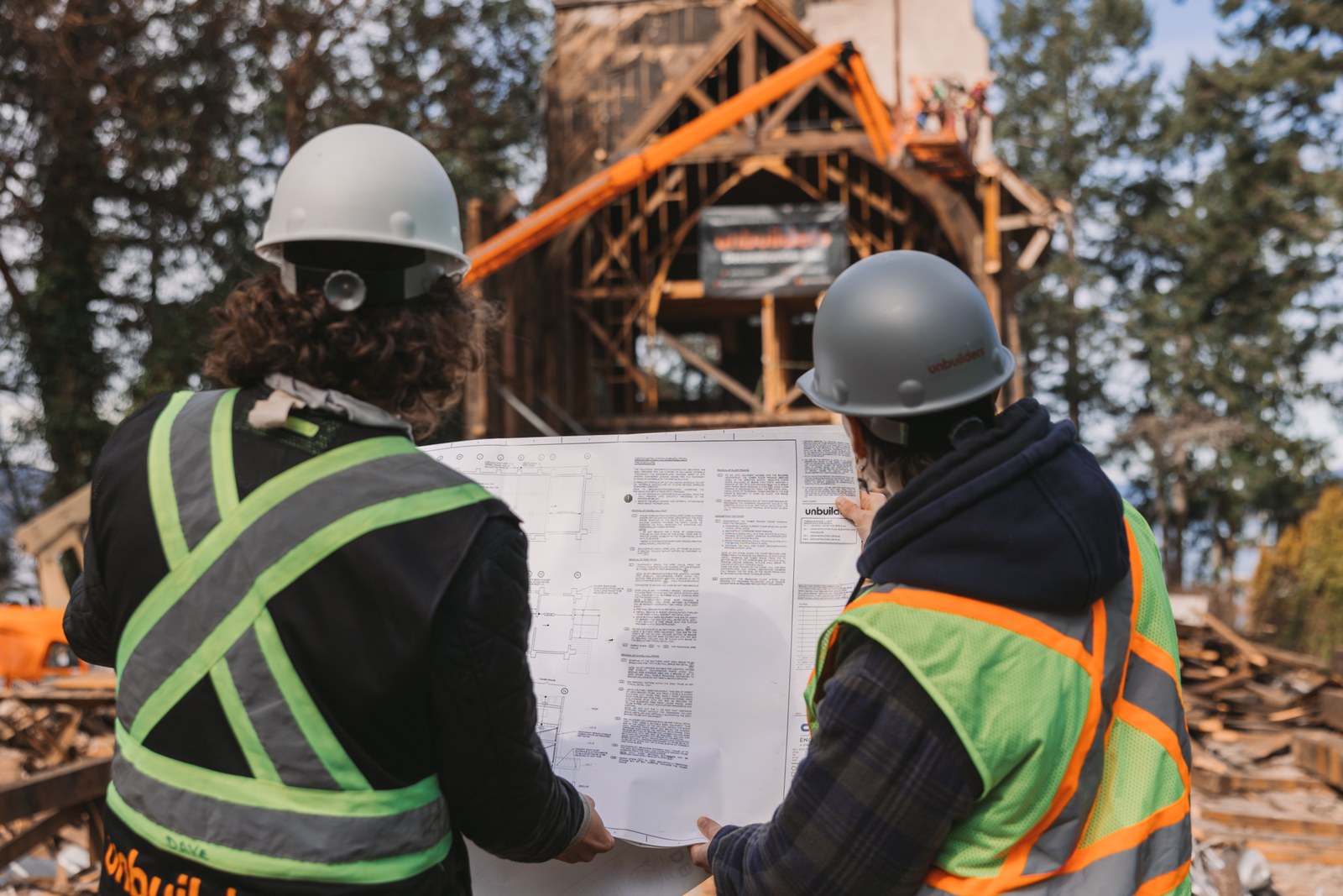 Two crew members review a large drawing with the exposed timber building behind them